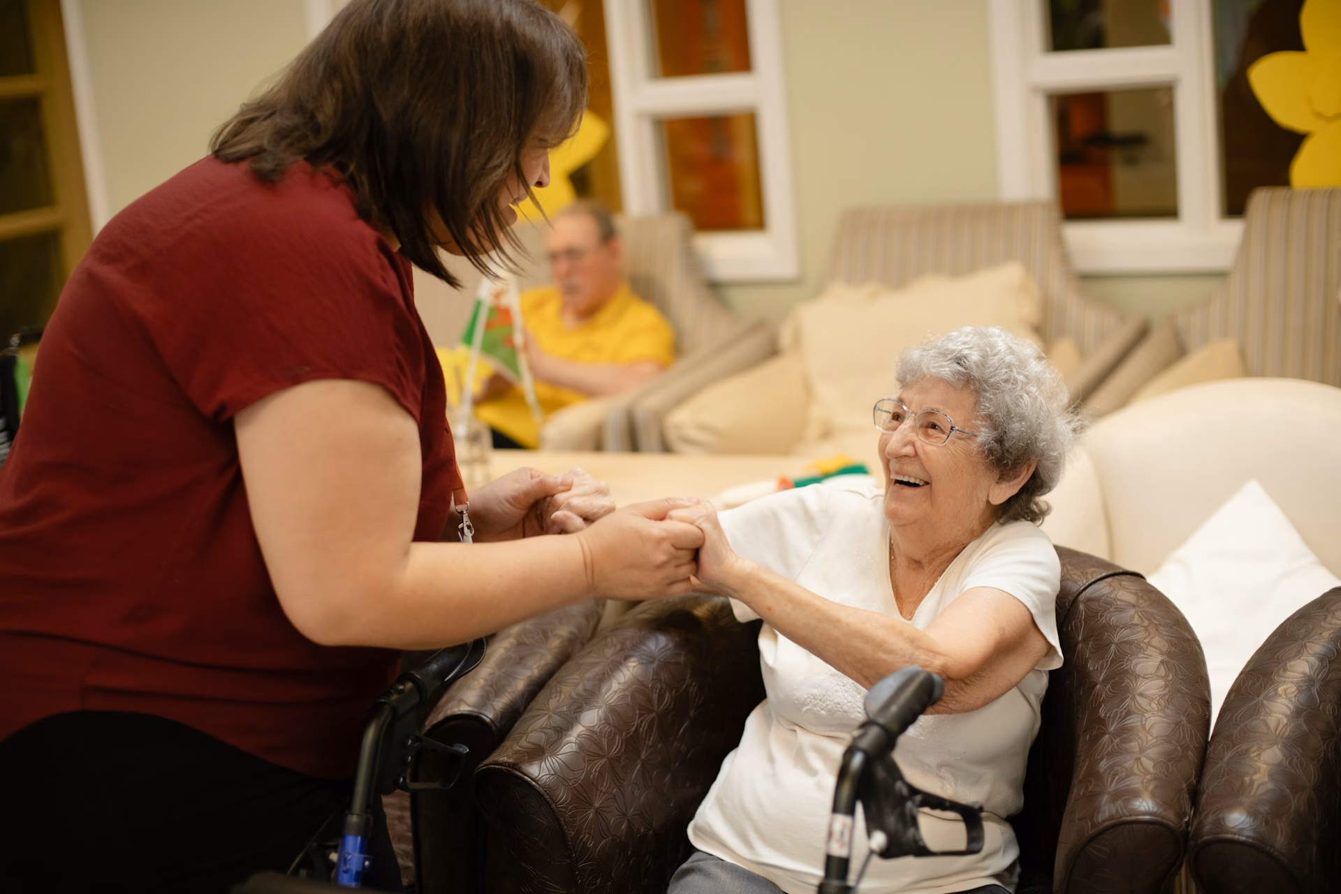 Older woman in chair smiling and holding hands with caregiver in a warm, homey room.