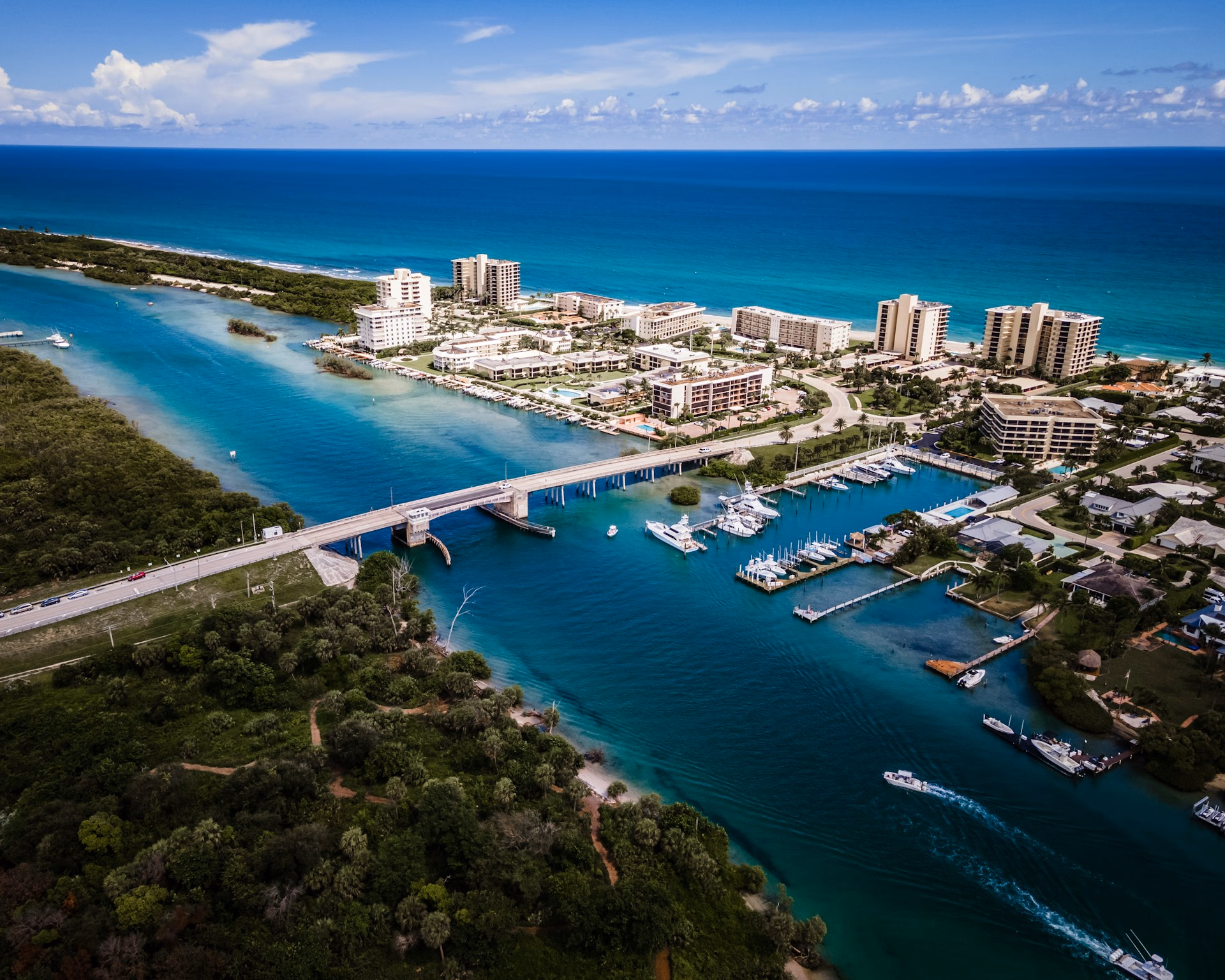 Aerial view of a coastal town with a bridge, boats, residential buildings, and blue ocean under a partly cloudy sky.