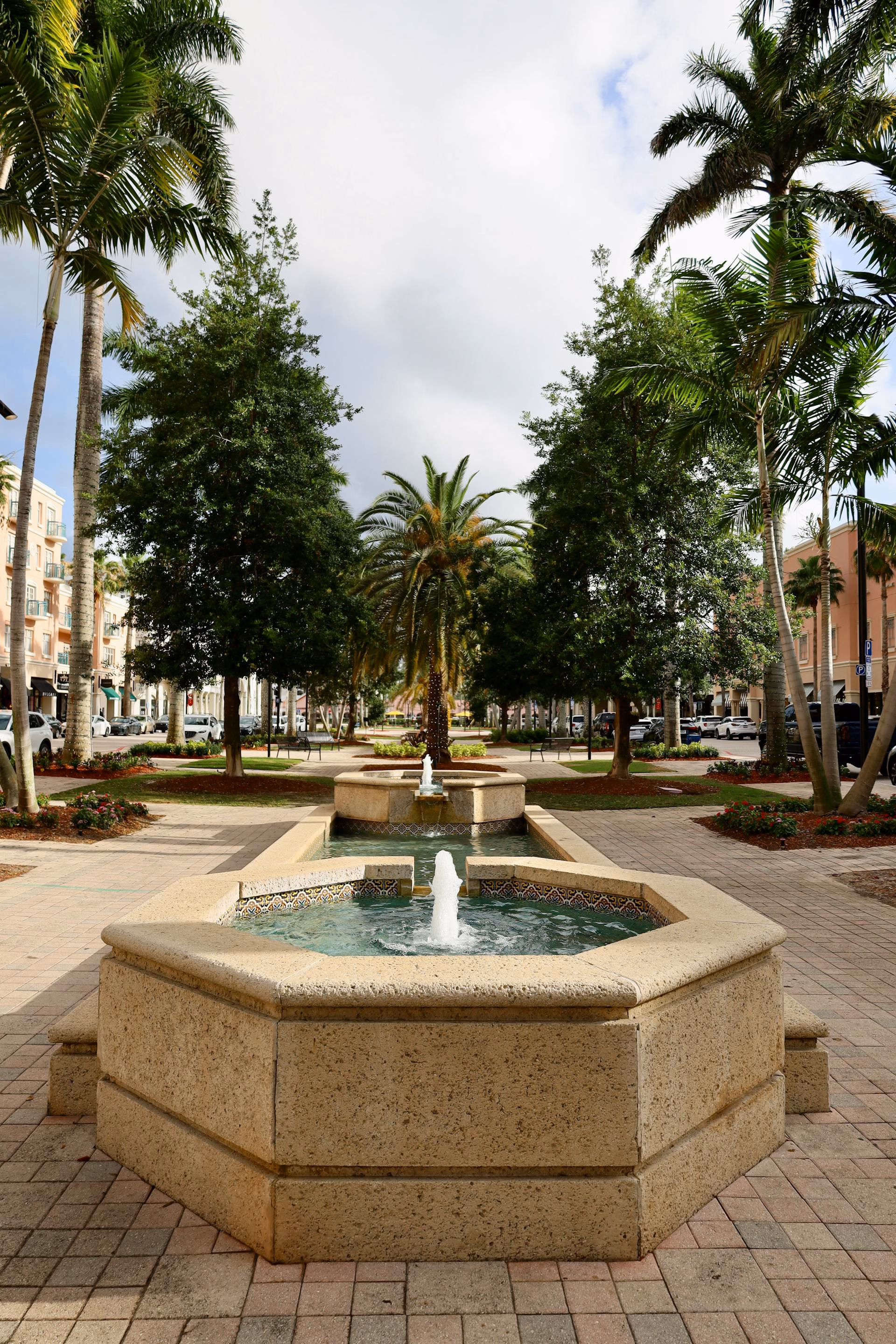 Two tiered hexagonal stone fountains along a tree-lined pedestrian walkway with palm trees.