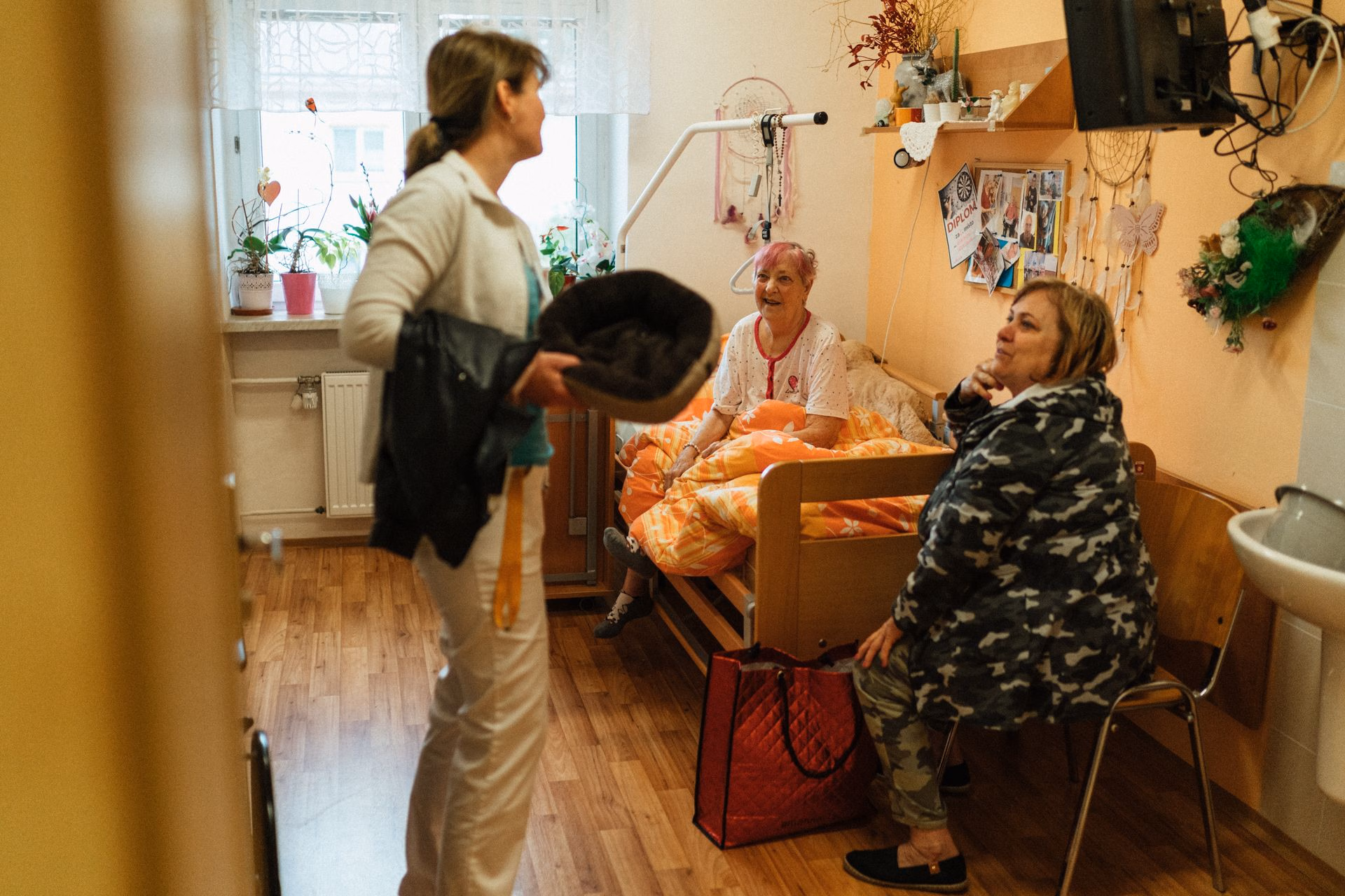 Three women in a cozy room, one sitting on a bed, one on a chair, and one standing with a bag and coat.