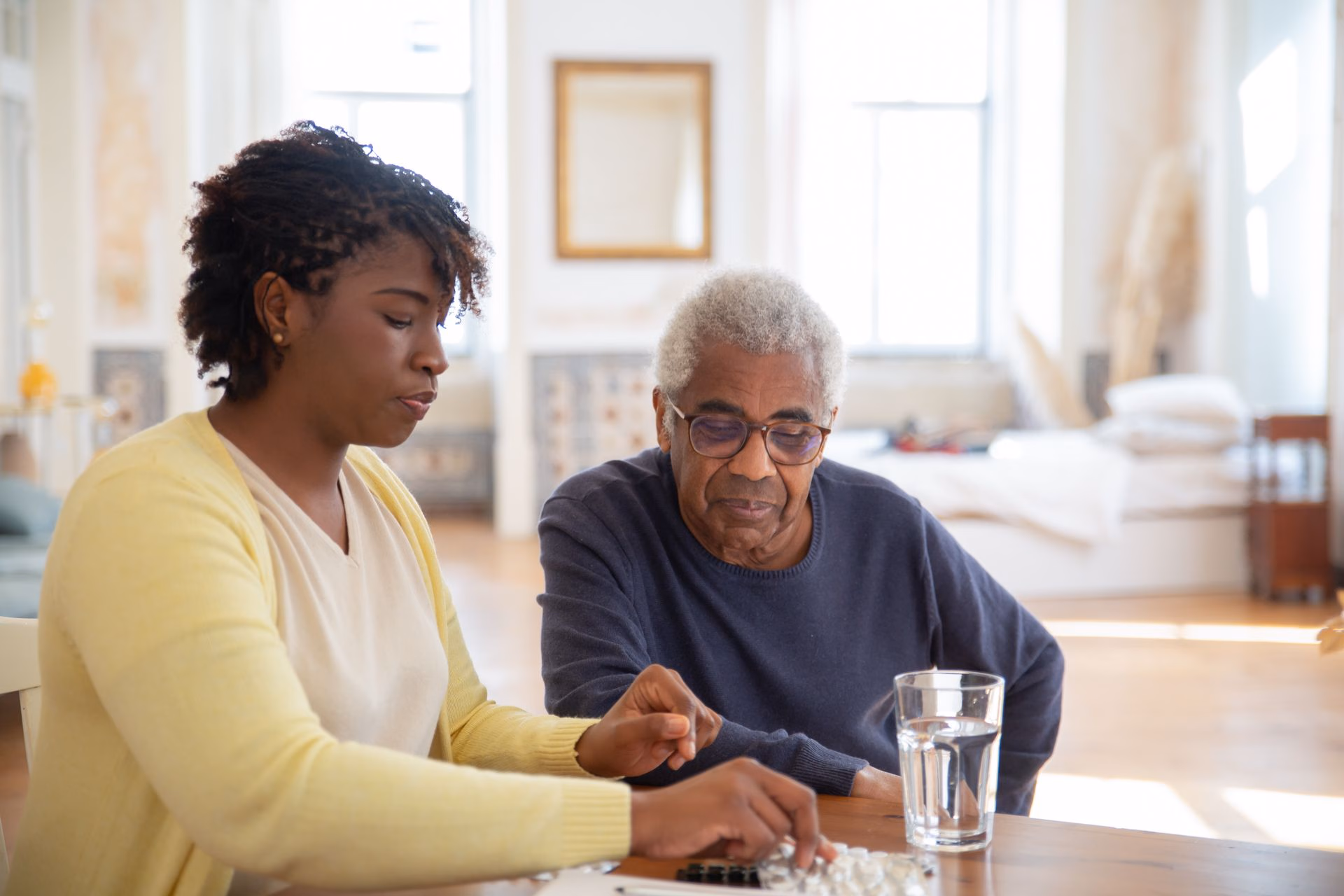 Woman helping elderly man with medication at a table with glass of water in a bright room.