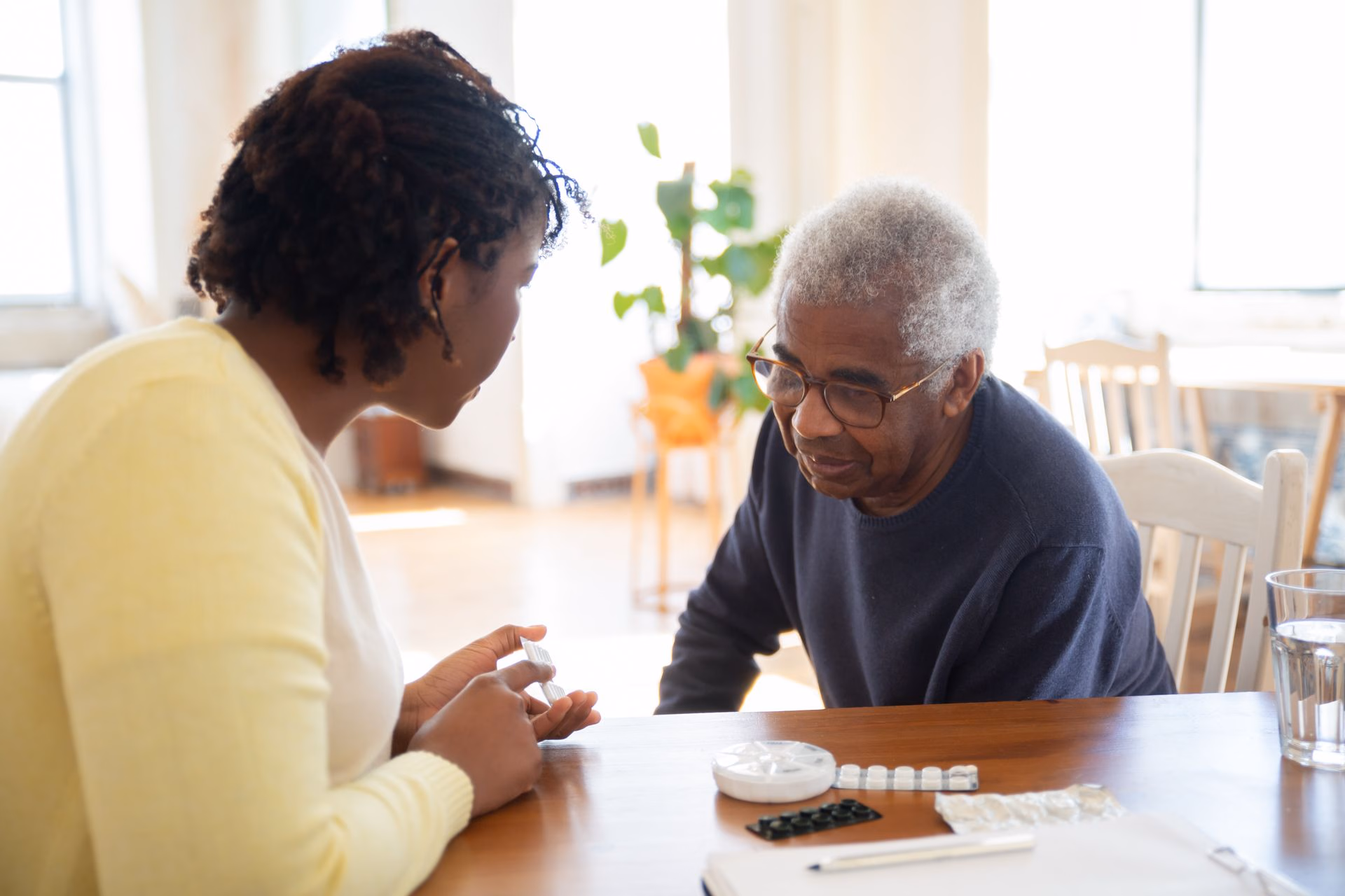Woman helping elderly man manage medications at a table with pills and a glass of water.
