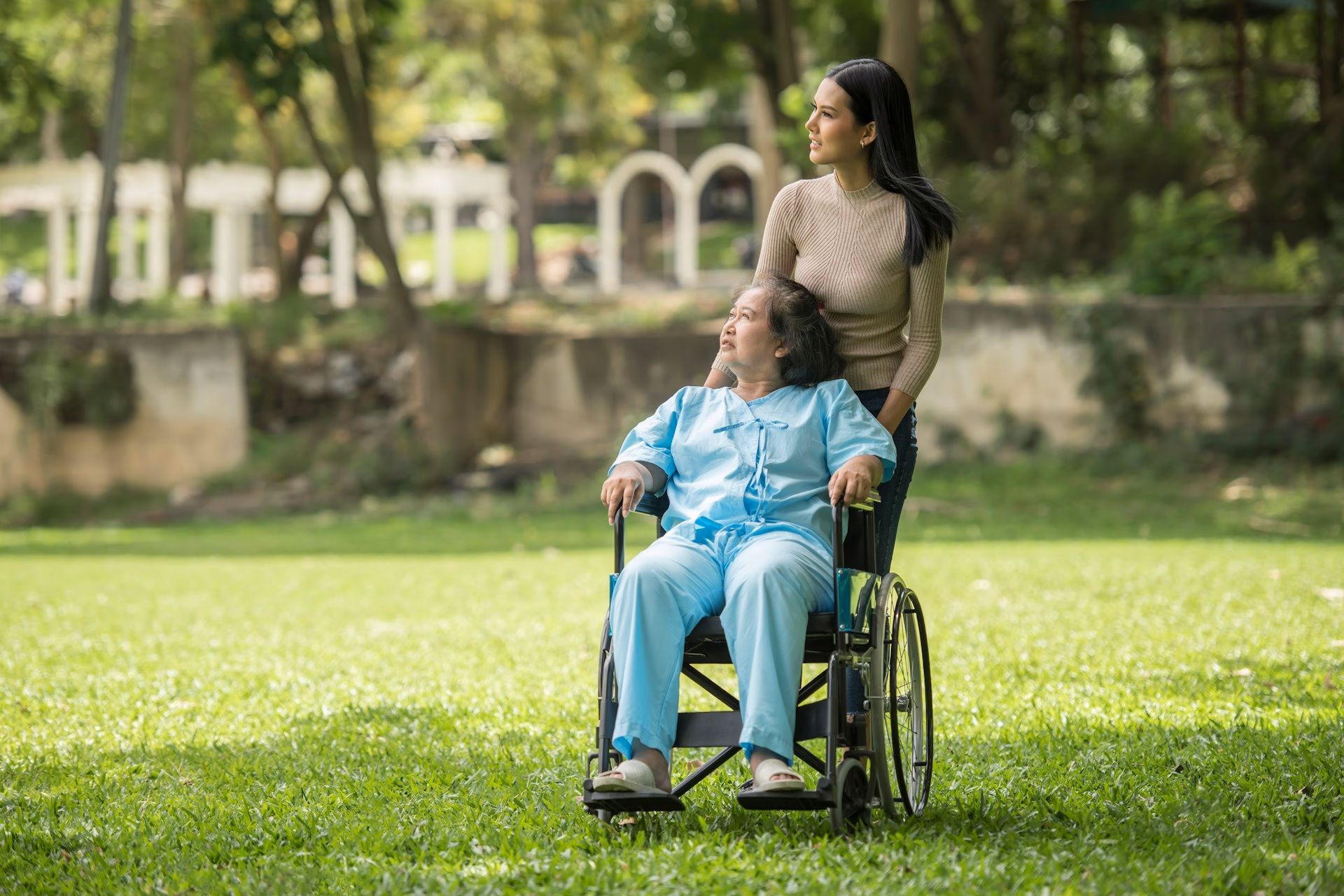 Young woman standing behind an elderly woman in a wheelchair in a green park looking to the right.