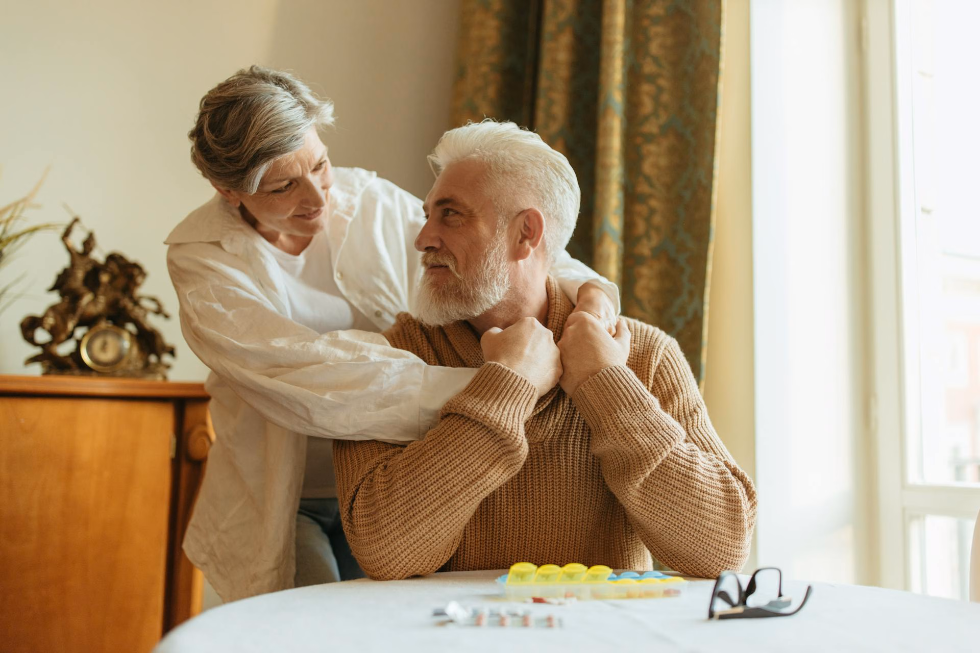 Elderly woman warmly embraces seated elderly man looking at her at a table with medication and glasses.