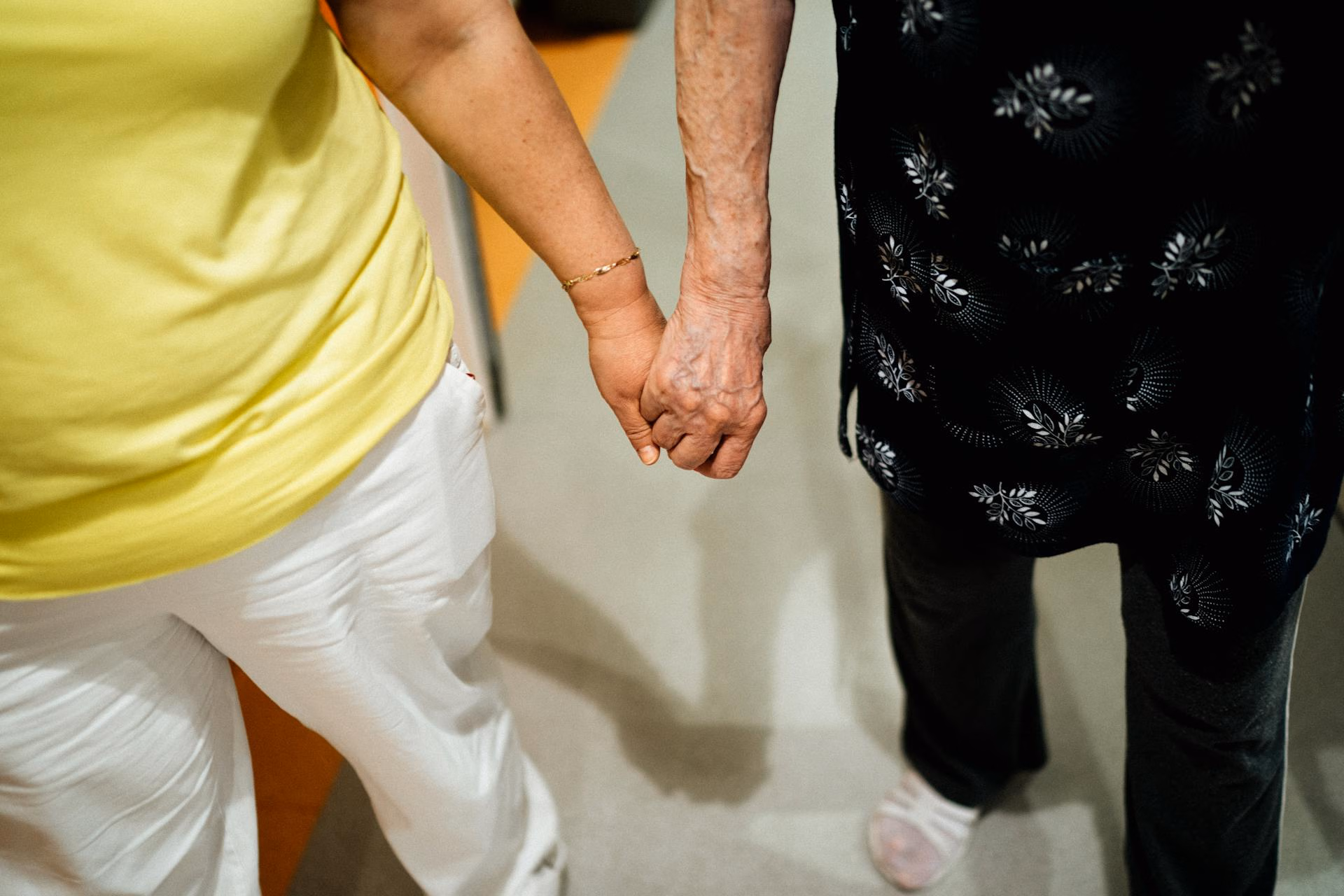 Close-up of two people holding hands, one wearing white pants and yellow shirt, the other black patterned dress.