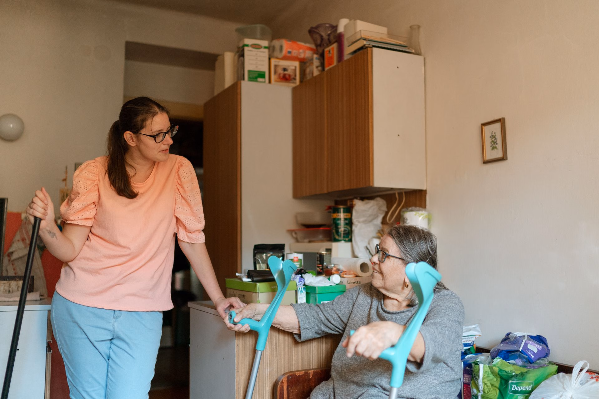 Young woman in peach top supports elderly woman with crutches in a cluttered room.