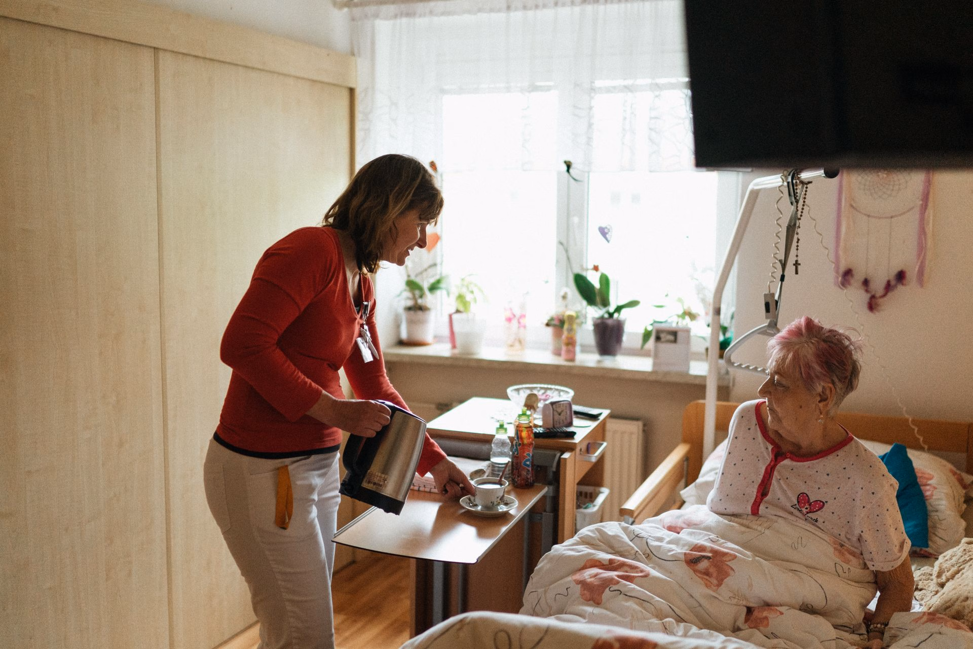 Caregiver in red shirt serving tea to elderly woman sitting in bed in cozy room with plants.