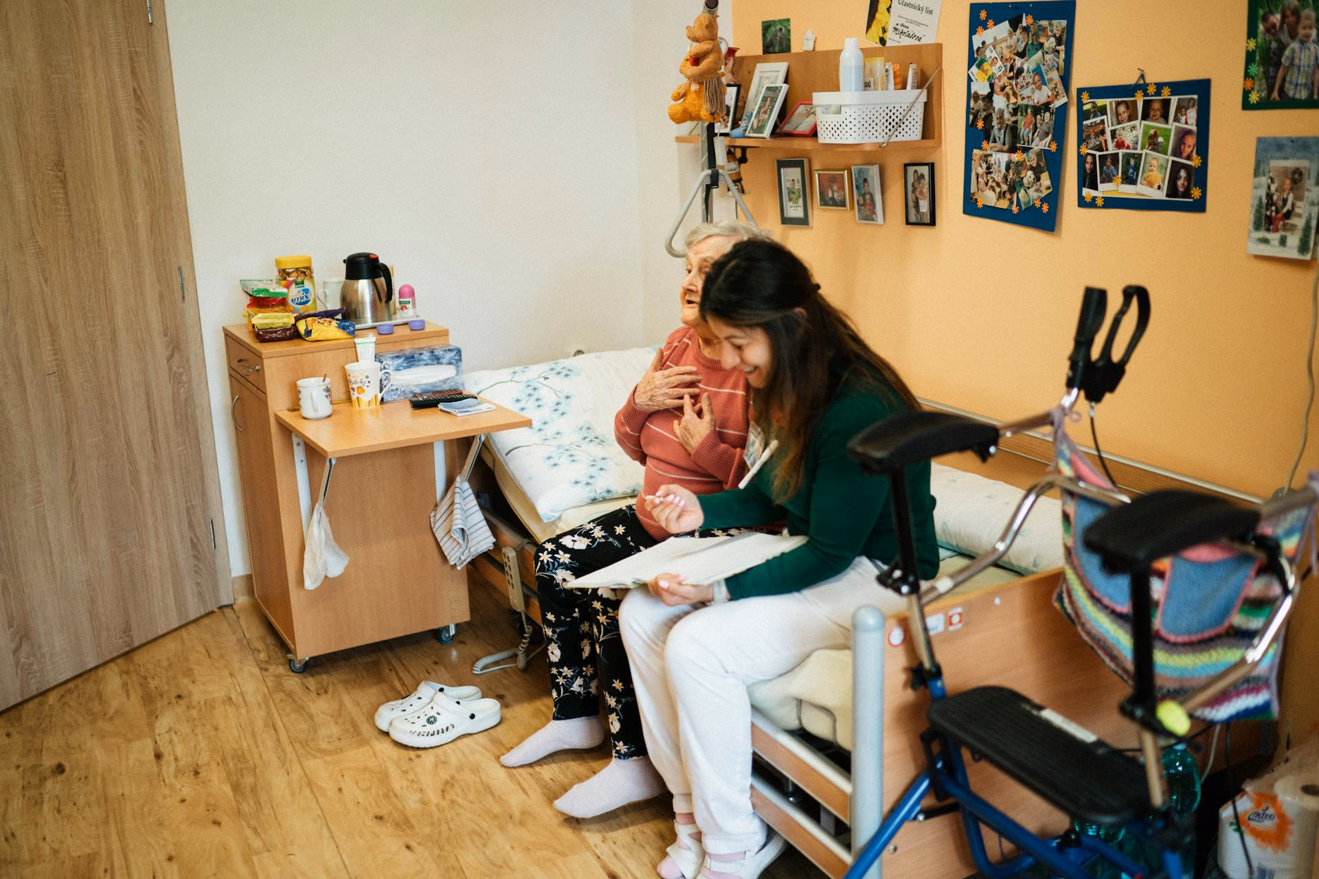 Elderly woman and caregiver sitting on bed, smiling and looking at clipboard in cozy room.